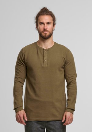 Mann mit Bart und zusammengebundenem Haar, der ein braunes Langarm-Henley-Shirt und graue Hosen trägt und vor einem einfarbigen Hintergrund steht.