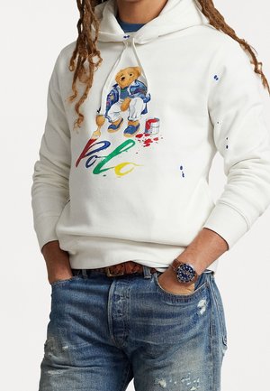 White hoodie with colorful bear painting "Polo" text on front, worn with distressed blue jeans and a wristwatch.