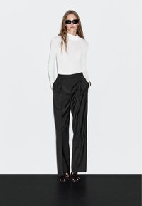 Witte lange mouw turtleneck, grijze high-waisted broek met plooien, zwarte platform sandalen en donkere zonnebril tegen een effen achtergrond.
