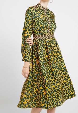 Femme portant une robe jaune à manches longues, longueur genou, avec un imprimé léopard vert et noir, et une ceinture et un col à motifs.