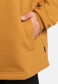 Sudadera amarilla mostaza con un bolsillo lateral, puños acanalados y una etiqueta negra en el lateral. Textura suave y corte relajado.