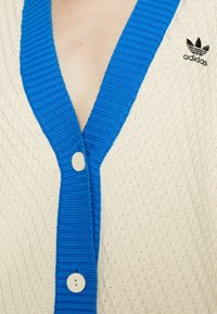 Krämfärgad cardigan med blå ribbad krage och knappar. Har Adidas-logotypen i svart på övre vänstra sidan.