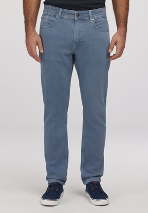 Mężczyzna w jasnoniebieskich dżinsach i granatowych sneakersach, stojący na tle gładkiego jasnoszarego tła.