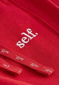 Red fabric featuring an embroidered white logo spelling "self." and textured labels showing the same text. Smooth and vibrant finish.