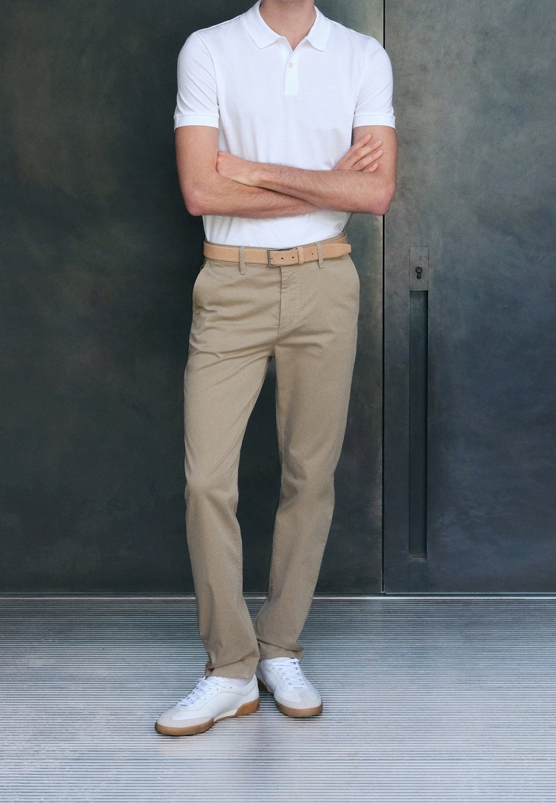 Homme debout, les bras croisés, portant un polo blanc, un pantalon beige avec une ceinture et des baskets blanches sur un fond métallique sombre.