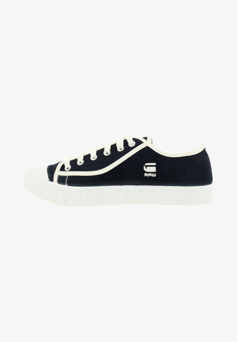 Zapatillas de lona de color marino con puntera de goma blanca y suela texturizada, que cuentan con cordones blancos y logotipo en el costado. Forma redondeada con un diseño de bajo perfil.