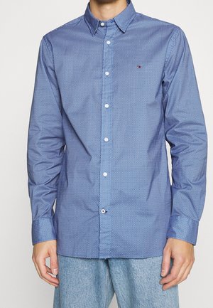 Mann trägt ein blau gemustertes Hemd mit Knöpfen und kleinem Tommy Hilfiger-Logo auf der Brust, kombiniert mit hellblauen Jeans.