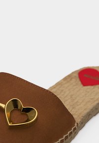 Καφέ σανδάλι με σαγιονάρα, με σόλα από τζούτι και χρυσό μεταλλικό κόσμημα σε σχήμα καρδιάς στο πάνω λουρί, πάνω σε απλό φόντο.