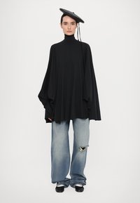 Hollarin musta kauluspaidan, jossa on leveät hihat, yhdistettynä vaaleansiniin revittyihin farkkuihin ja mustiin kenkiin. Musta hattu on asetettu vinosti.