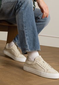 Sneakers beige in suede con superficie texturizzata, suola in gomma bianca e punta rotonda. Indossati con calze bianche e jeans azzurri chiari.