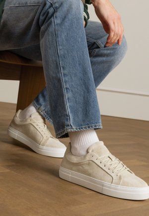 Beige suède sneakers met een gestructureerd oppervlak, witte rubberen zool, en ronde neus. Gedragen met witte sokken en lichtblauwe jeans.