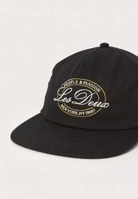 Fekete baseballsapka lapos karimával, fehér és sárga hímzett logóval, amelyen a "Les Deux" és a "People & Passion" feliratok olvashatók.