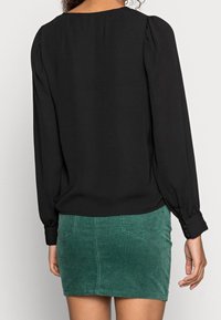 Blouse noire à manches longues avec épaules bouffantes, associée à une mini-jupe en velours côtelé verte avec poches et surface texturée.