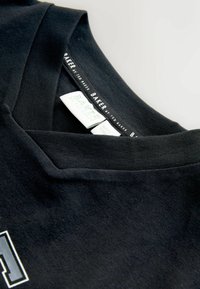 Czarny bawełniany t-shirt z dekoltem w kształcie litery V. Metki są widoczne przy dekolcie, wyświetlając nazwę marki na białym tle. Gładka tekstura.