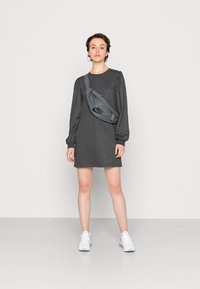 Vestido cinza de mangas longas com ombros volumosos, combinado com uma mochila cinza da Nike com um logotipo preto. Modelo a usar ténis brancos.