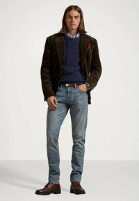 Man med mörkbrun kavaj, blå kabelstickad tröja, ljusblå jeans, bruna läderskor och klocka, stående mot en neutral bakgrund.