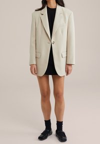 Beige oversized kavaj med två framfickor, enkel knappstängning och strukturerade axlar, bärs över en svart klänning och svarta skor.