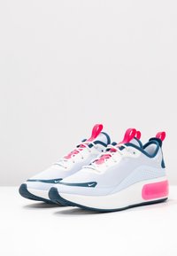 Pár bílých a modrých atletických tenisek s růžovými akcenty, tkaničkami a patními taby vystavený na bílé ploše na pozadí.