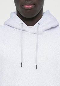 Ανοιχτό γκρι φούτερ με υφή υφάσματος, που διαθέτει κουκούλα με κορδόνι και κορδόνια με μεταλλικές άκρες. Απλός και καθημερινός σχεδιασμός.