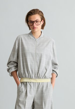 Junge Person mit kurzen Haaren und Brille trägt eine hellgraue karierten Reißverschlussjacke und passende Hose, Hände in den Taschen, neutraler Hintergrund.