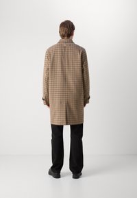 Braun karierter Überrock mit maßgeschneidertem Design, geknöpften Manschetten und gerader Silhouette. Getragen über schwarzen Hosen und Schuhen.