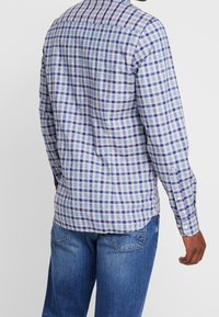 Camicia a maniche lunghe a quadri in blu, bianco e viola. Materiale di cotone, colletto con bottoni e orlo curvo. Indossata con jeans blu.