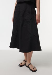 Falda negra de largo a la rodilla con patrones de bordado detallados, una textura suave y una silueta en línea A. La lleva puesta con sencillas chanclas negras.