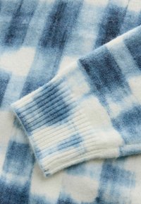 Pull en maille texturée présentant des nuances de bleu et de blanc dans un motif tie-dye, avec des poignets côtelés et un matériau doux et pelucheux.