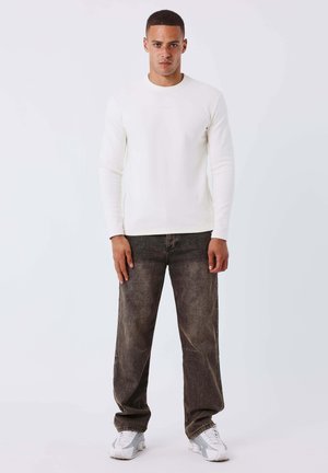 Homme debout faisant face, portant une chemise blanche à manches longues, un jean noir délavé et des baskets blanches, contre un fond clair uni.