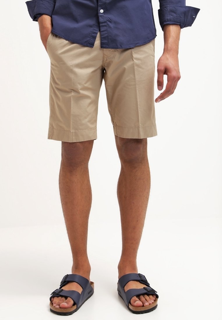 Homme portant un short beige jusqu'aux genoux, une chemise bleue marine à boutons et des sandales à boucle bleue marine, se tenant devant un fond blanc.
