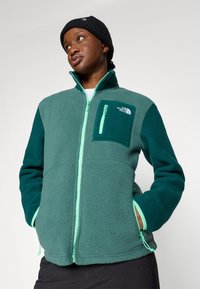 Fleece-jakke i mørk grønn og teal med en glidelåslomme på brystet. Har mintgrønn glidelås og mansjettakenter. Myk, teksturert stoff.