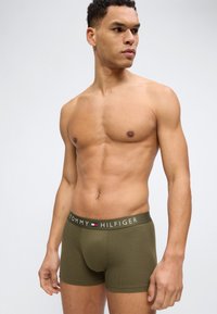 Muskelöser Mann trägt olivgrüne Tommy Hilfiger Boxershorts und steht mit seitlich gedrehtem Kopf vor einem schlichten hellen Hintergrund.