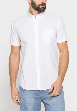 Uomo che indossa una camicia bianca a maniche corte con taschino sul petto e jeans azzurri, rivolto leggermente verso sinistra.