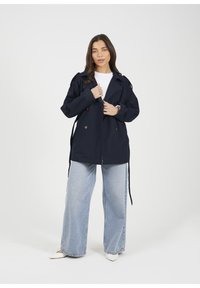 Granatowy płaszcz trench z podwójnymi guzikami i paskiem w talii, noszony na białej bluzce i w połączeniu z jasnoniebieskimi, szerokimi spodniami.