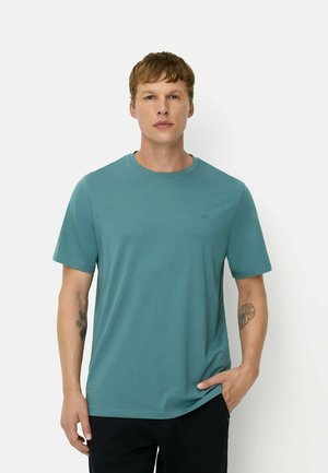 Mand iført turkis kortærmet t-shirt og sorte bukser, stående med den ene hånd i lommen mod en ensfarvet hvid baggrund.