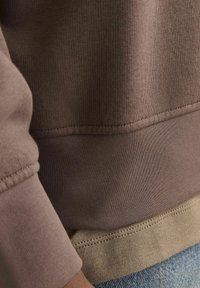 Ruskea huppari, jossa on pehmeä pinta, ribbikudotut hihansuut ja kerroksellinen ilme, jossa vaaleanruskea aluskerros näkyy helmassa.