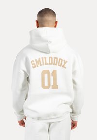 Felpa con cappuccio bianca con grande testo beige "SMILODOX" e "01" sul retro. Tessuto morbido, vestibilità rilassata e cappuccio regolabile.