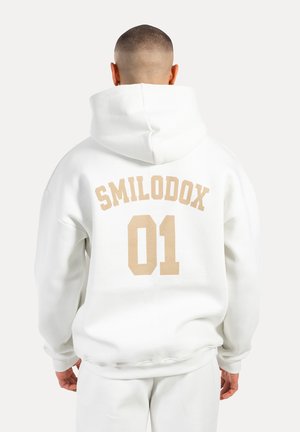 Sudadera blanca con gran texto beige "SMILODOX" y "01" en la parte de atrás. Tejido suave, corte holgado y capucha ajustable.