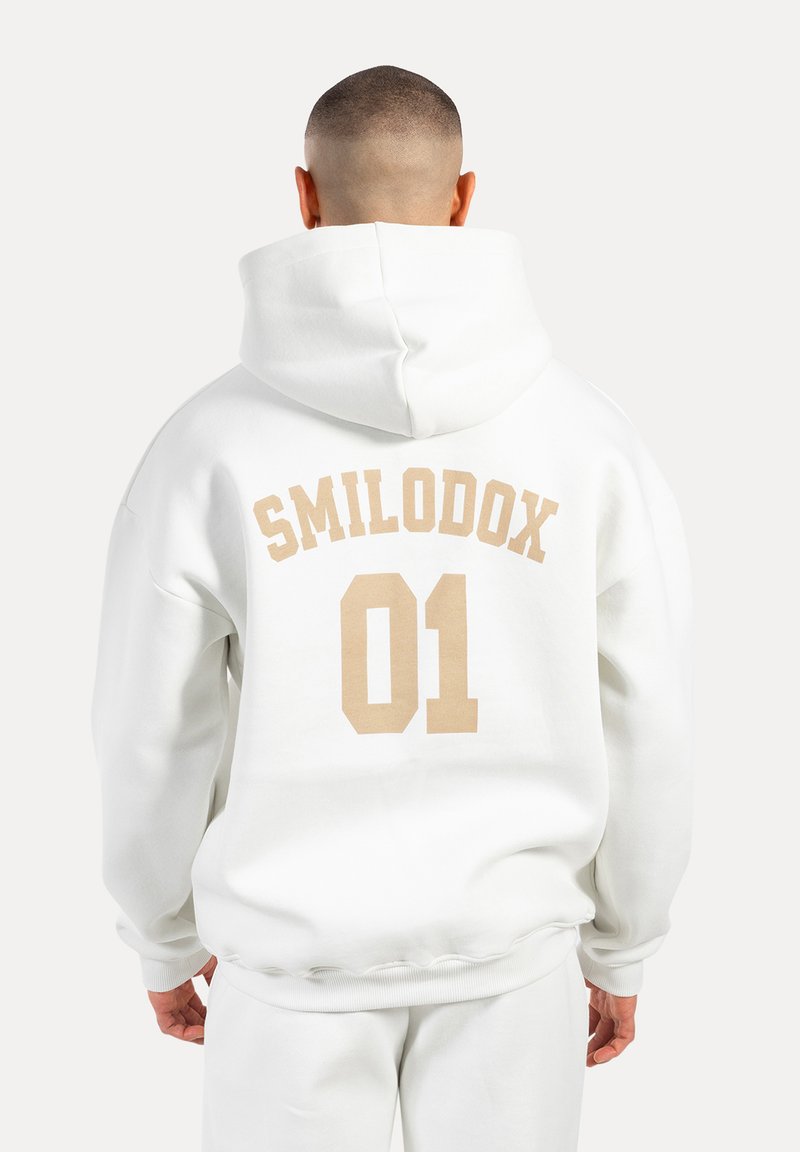 Felpa con cappuccio bianca con grande testo beige "SMILODOX" e "01" sul retro. Tessuto morbido, vestibilità rilassata e cappuccio regolabile.