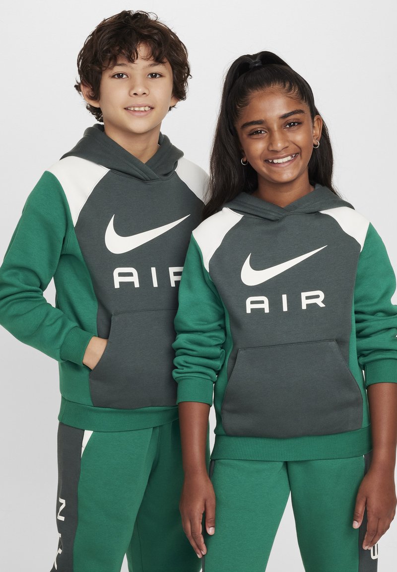 Dwoje dzieci nosi dopasowane bluzy Nike w ciemnozielonym i szarym kolorze, z dużym białym logo i kieszeniami z przodu. Bluzy mają ściągacze na mankietach.