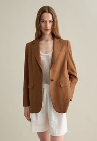 Blazer in lino marrone con due tasche anteriori, chiusura con un bottone e rever a taglio, abbinato a un top bianco testurizzato e pantaloni corti.