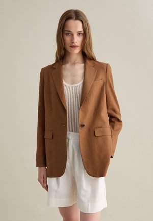 Blazer de lino marrón con dos bolsillos frontales, cierre de un botón y solapas con muesca, combinado con una blusa blanca texturizada y pantalones cortos.
