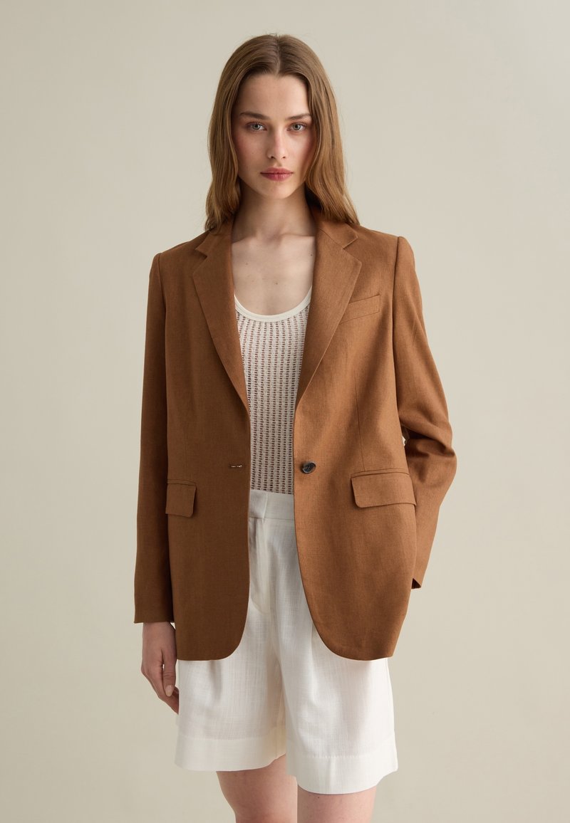 Blazer in lino marrone con due tasche anteriori, chiusura con un bottone e rever a taglio, abbinato a un top bianco testurizzato e pantaloni corti.