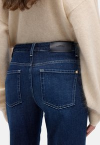 Jeans di denim blu scuro con un'etichetta in pelle sul retro, caratterizzati da due tasche posteriori e una texture liscia, abbinati a un maglione beige.