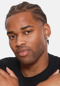 Pendientes plateados y negros con un diseño geométrico, colgando de un arete, llevados por una persona con cabello trenzado, sobre un fondo blanco.
