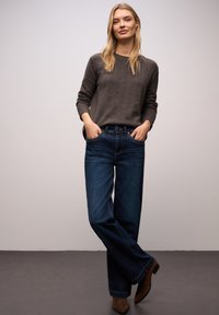 Dunkelgrauer Strickpullover und dunkelblaue Jeans mit weitem Bein, ausgestattet mit Fronttaschen und braunen Stiefeletten. Das Modell steht mit den Händen in den Taschen.