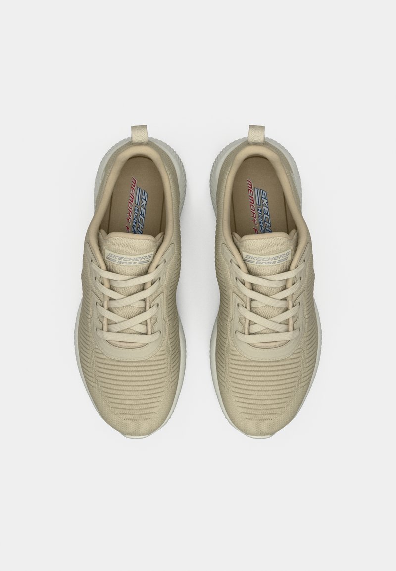 Zapatillas deportivas beige con parte superior texturizada, punta redonda y suela de goma. Las características incluyen cordones tonales y la marca en la lengüeta.