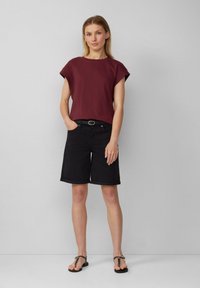 Burgunderfarbenes kurzärmeliges Baumwolloberteil kombiniert mit schwarzen, knielangen Denim-Shorts, die einen umgeschlagenen Saum und Fronttaschen aufweisen. Schwarze Flip-Flops.