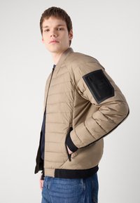 Casaco bomber isolado beige com bolsos laterais com fecho, punhos canelados e um detalhe preto no bolso da manga esquerda. Textura suave e design volumoso.