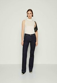 Eine Frau trägt ein weißes ärmelloses Top, dunkelblaue Jeans, schwarze spitze Schuhe und hält eine kleine schwarze Umhängetasche, während sie vor einem schlichten Hintergrund steht.